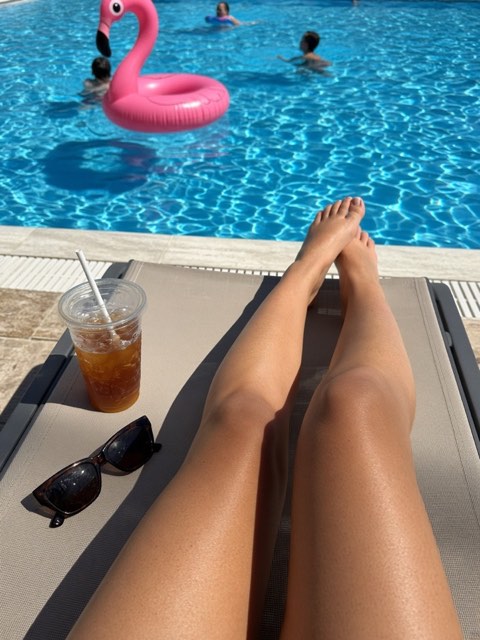 legs stretched out by a pool with a pink flamingo float and an iced drink
